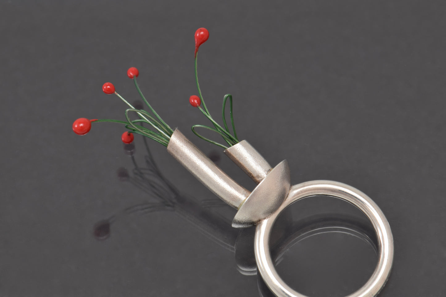 Silver ring with a small vase containing red buds on green stems on a dark background