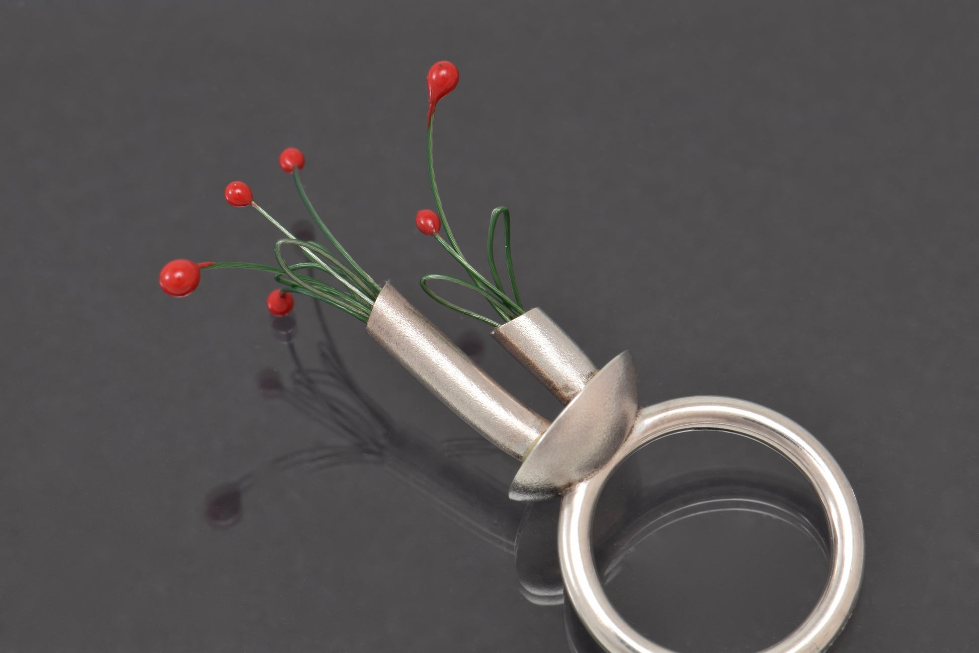 Silver ring with a small vase containing red buds on green stems on a dark background